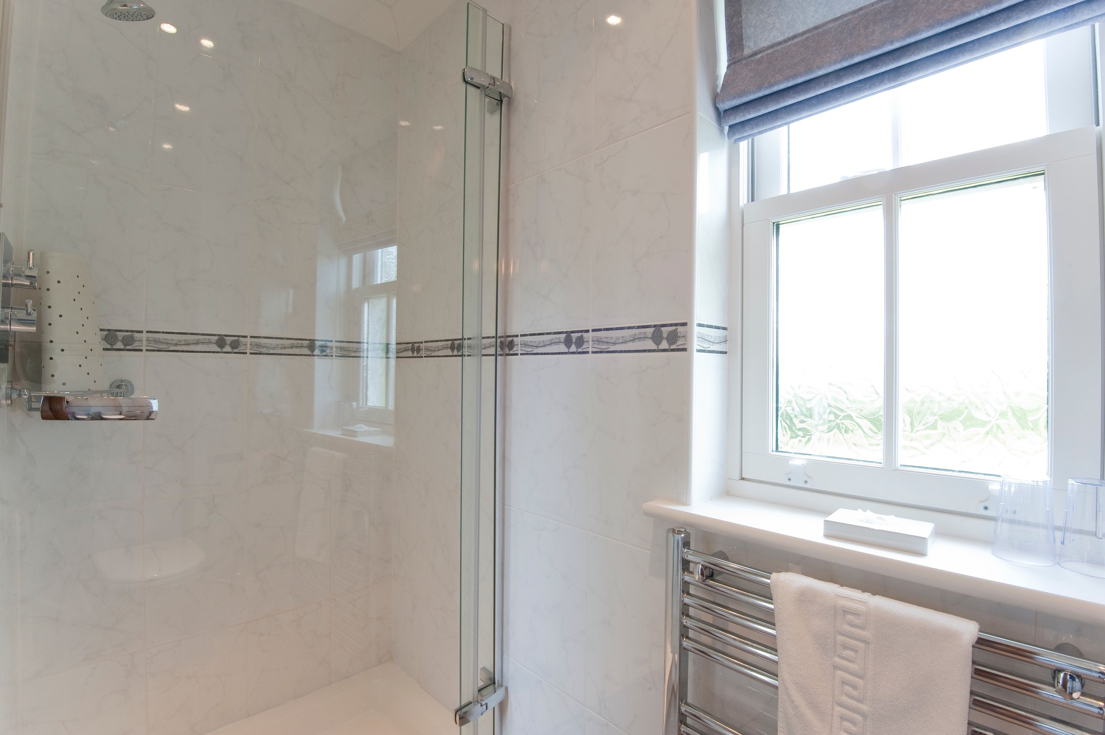 Ein modernes Badezimmer mit einer Glaskabinedusche, einem Waschtisch mit Handtuchhalter und einem Fenster mit Blick auf Grünflächen.