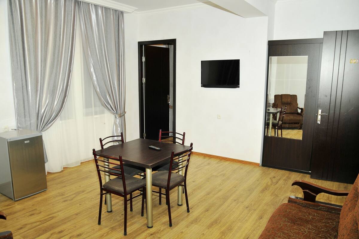 A dining area with a wooden table and chairs, a television on the wall, and a door with a number.