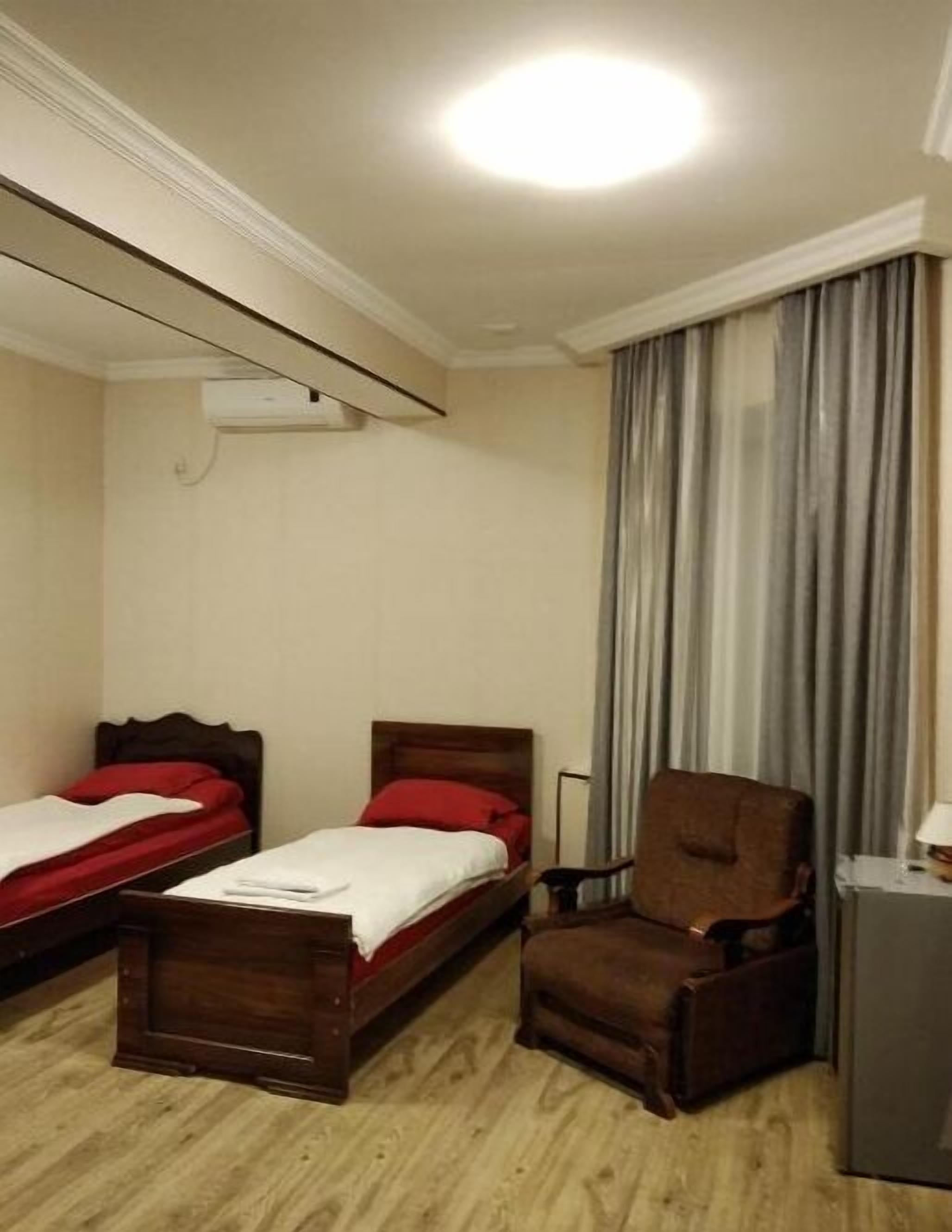 Een hotelkamer met twee eenpersoonsbedden, een houten stoel, een bureau met een lamp en een groot raam met gordijnen.