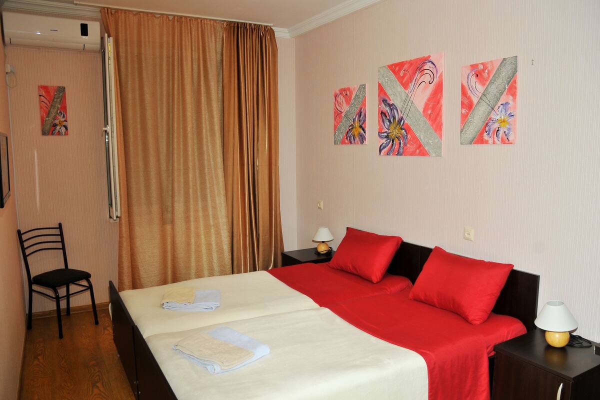 A hotel room with a double bed, red bedding, two nightstands with lamps, a chair, and artwork on the wall.