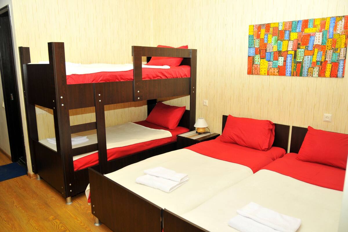 A room with bunk beds and a single bed, both with red bedding. There is a small table with a lamp and a colorful artwork on the wall.
