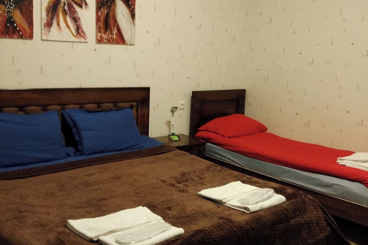 A bedroom with two beds, a large bed with a brown cover and two smaller beds with red and blue covers. There are three paintings on the wall above the beds.