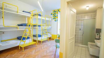 A dormitory room with bunk beds, a wooden floor, a chair, and a bathroom with a shower and toilet.