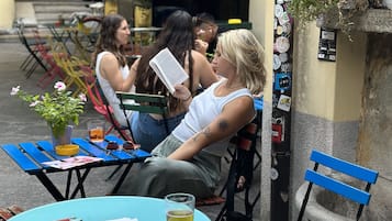 A woman reading a book at an outdoor café with a table, chairs, and a potted plant.