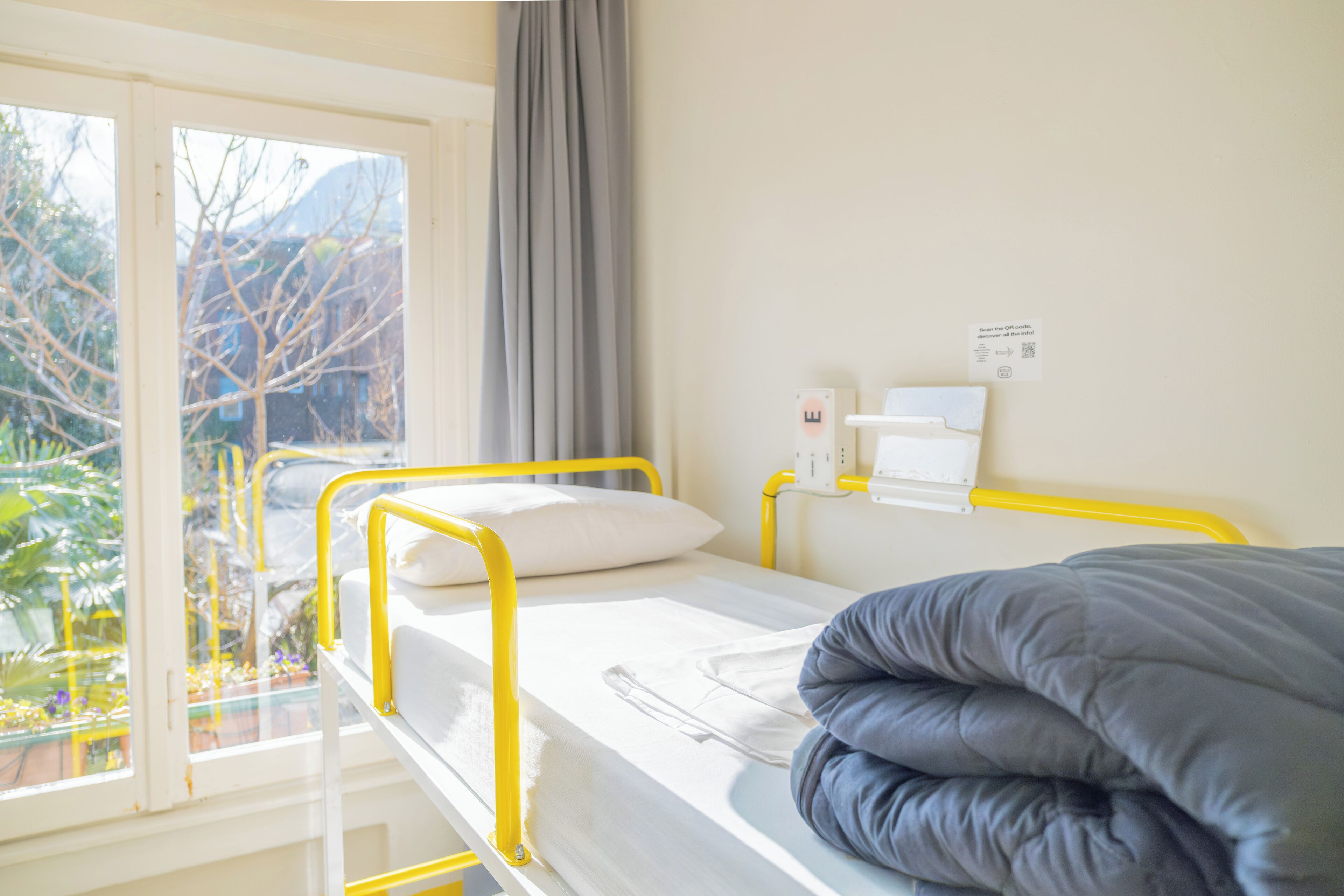 A single-bed room with a window, a curtain, a bedside table, and a wall-mounted device.
