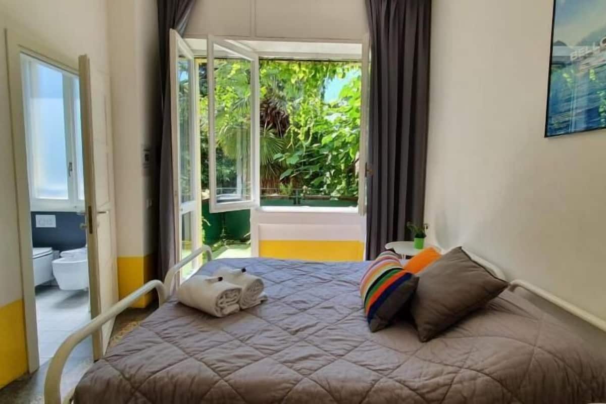 A bedroom with a bed, a bedside table, a window, and a view of greenery outside.
