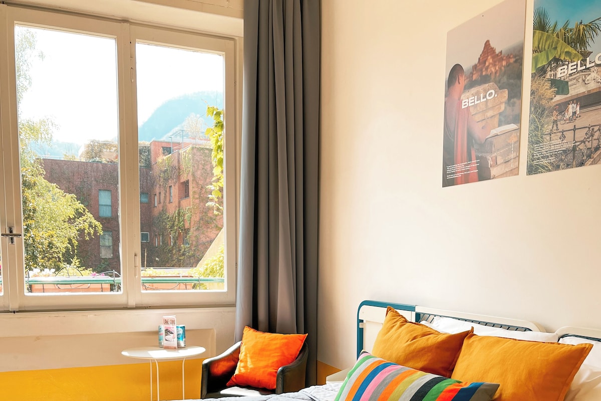 A hotel room with a bed, pillows, a small table, and a window with curtains.