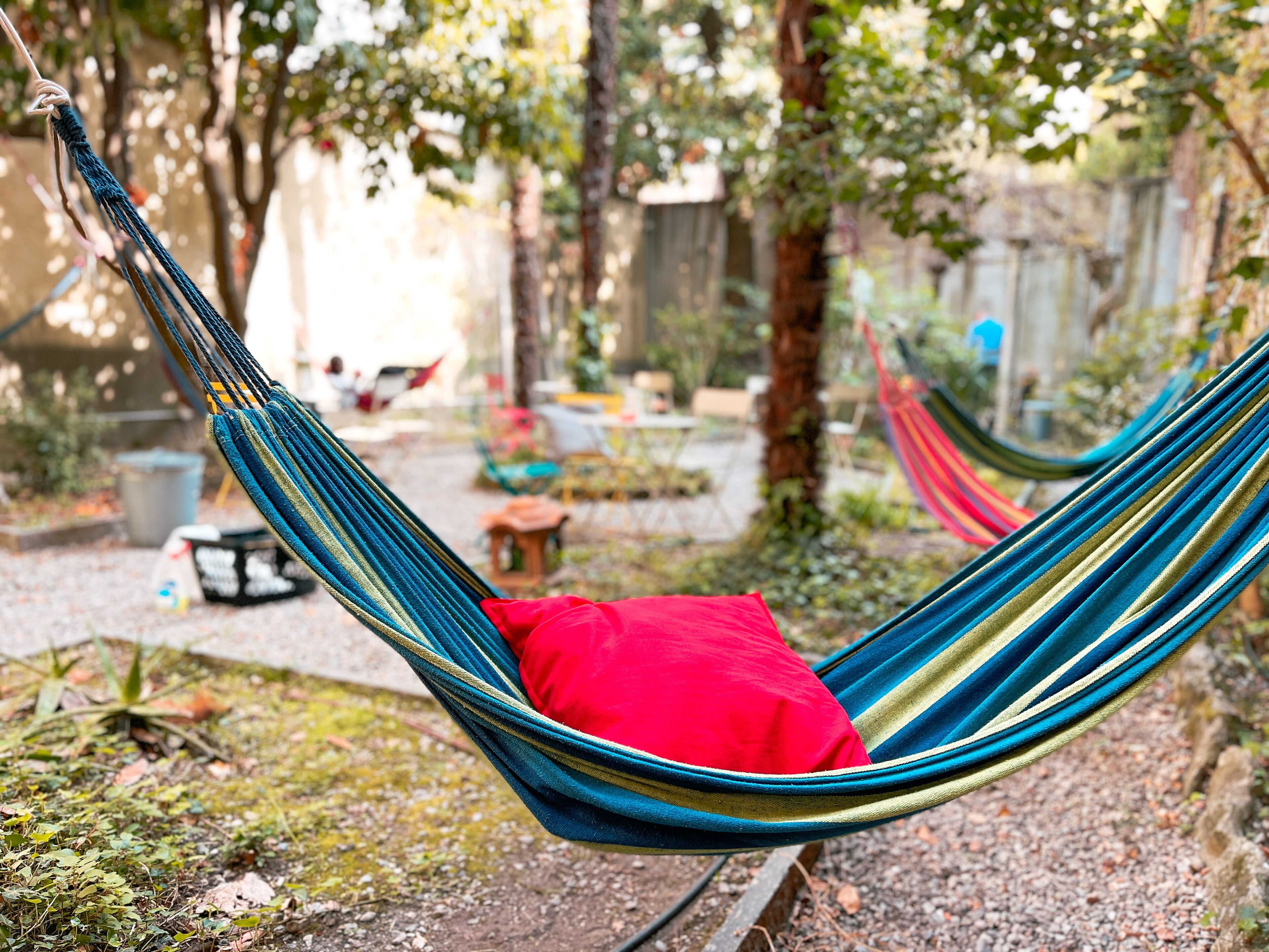 A hammock in a garden with a red pillow, surrounded by trees and plants.