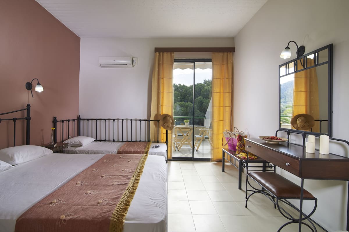 Habitación de hotel con dos camas, un escritorio con espejo, un banco y un balcón con cortinas amarillas.
