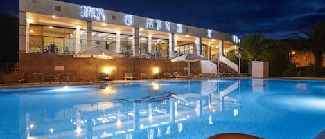 A swimming pool with lounge chairs and umbrellas, a lifebuoy, and a building with illuminated signage.