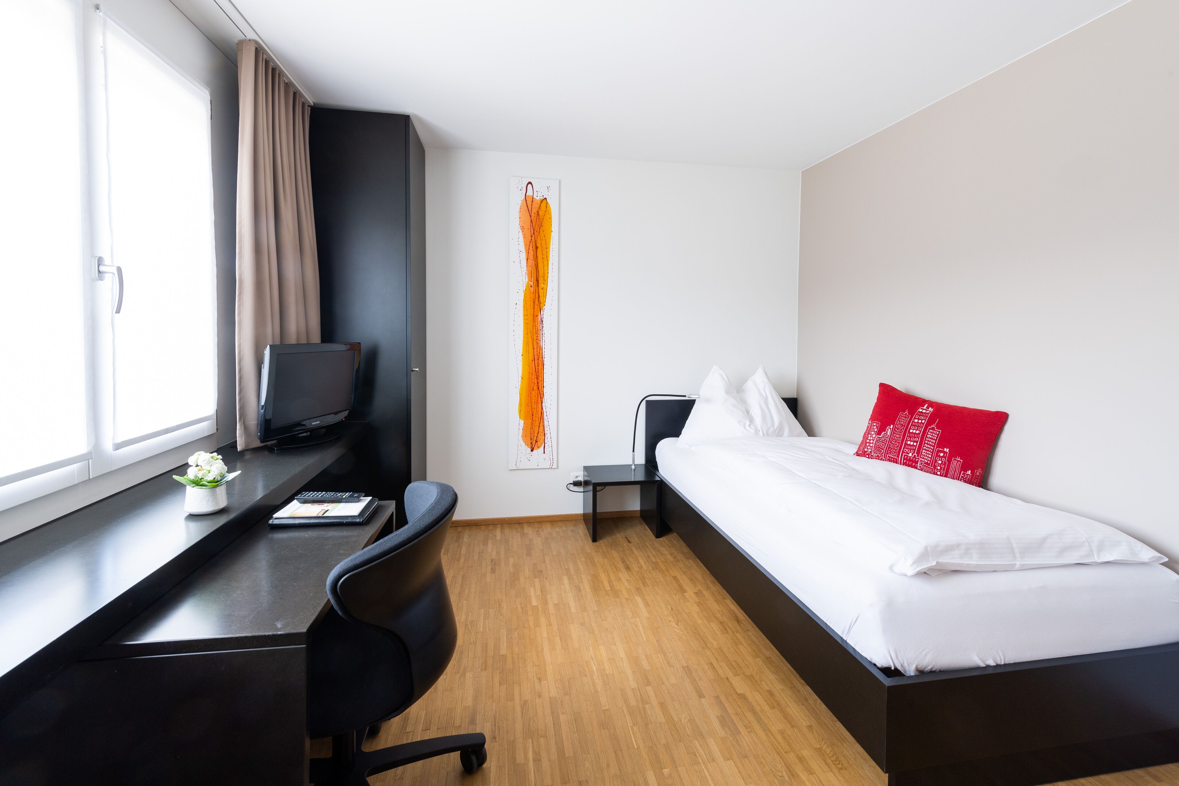 A modern hotel room with a wooden floor, a bed with white bedding and a red pillow, a desk with a computer and a small plant, and a large window with curtains.