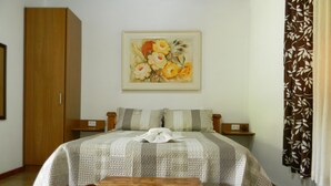 A bedroom with a wooden bed, a quilted bedspread, a wooden bench, a painting of flowers on the wall, and a brown patterned curtain. - Chalés Luar da Serra (Santo Antonio do Pinhal)