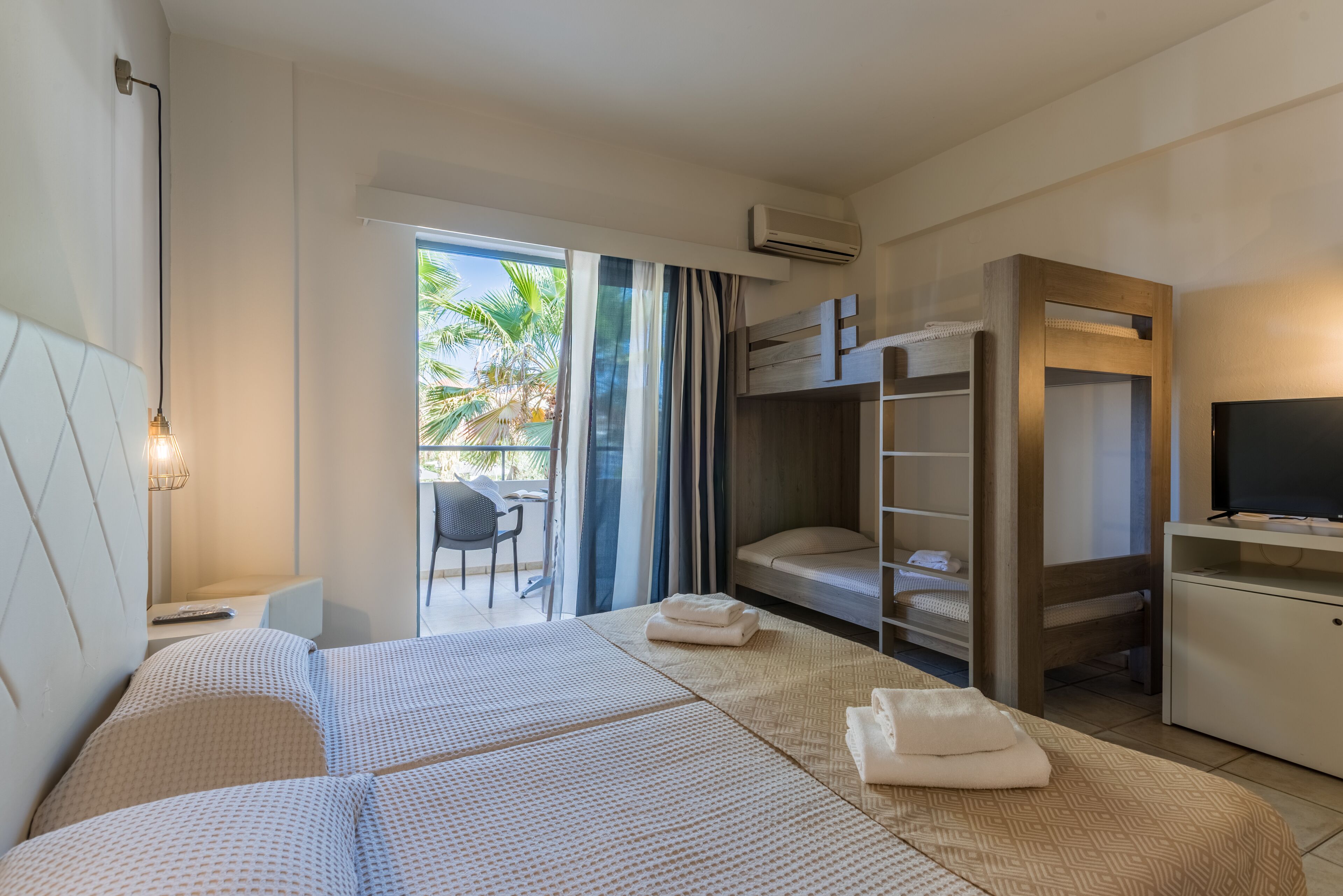 Habitación de hotel con cama, escritorio, televisor, literas y balcón con vista a palmeras.