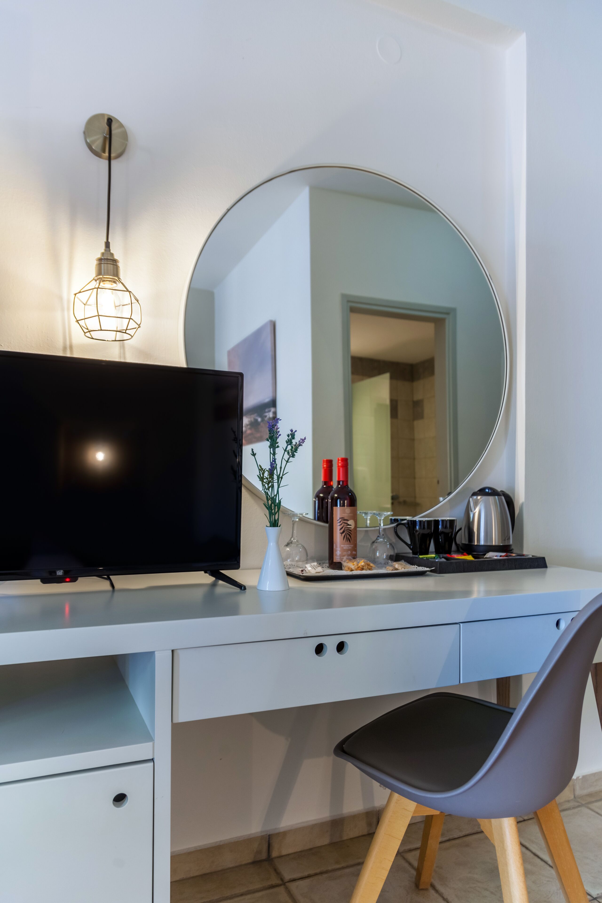 A modern hotel room with a flat-screen TV, a round mirror, a desk with a chair, a small vase with flowers, and a coffee maker.