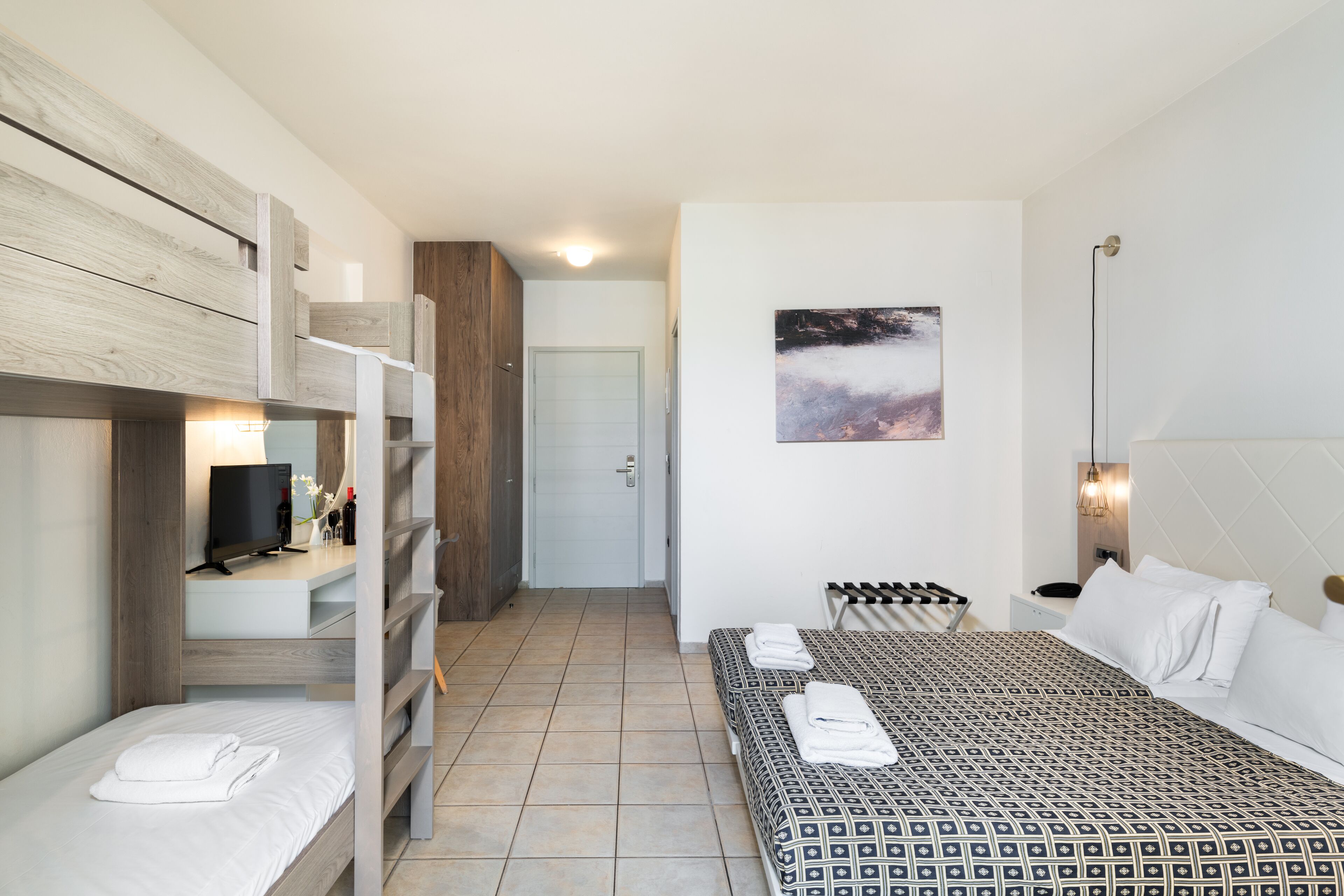 Quarto de hotel com beliche, uma cama de solteiro, televisão, escrivaninha com computador e um quadro na parede.
