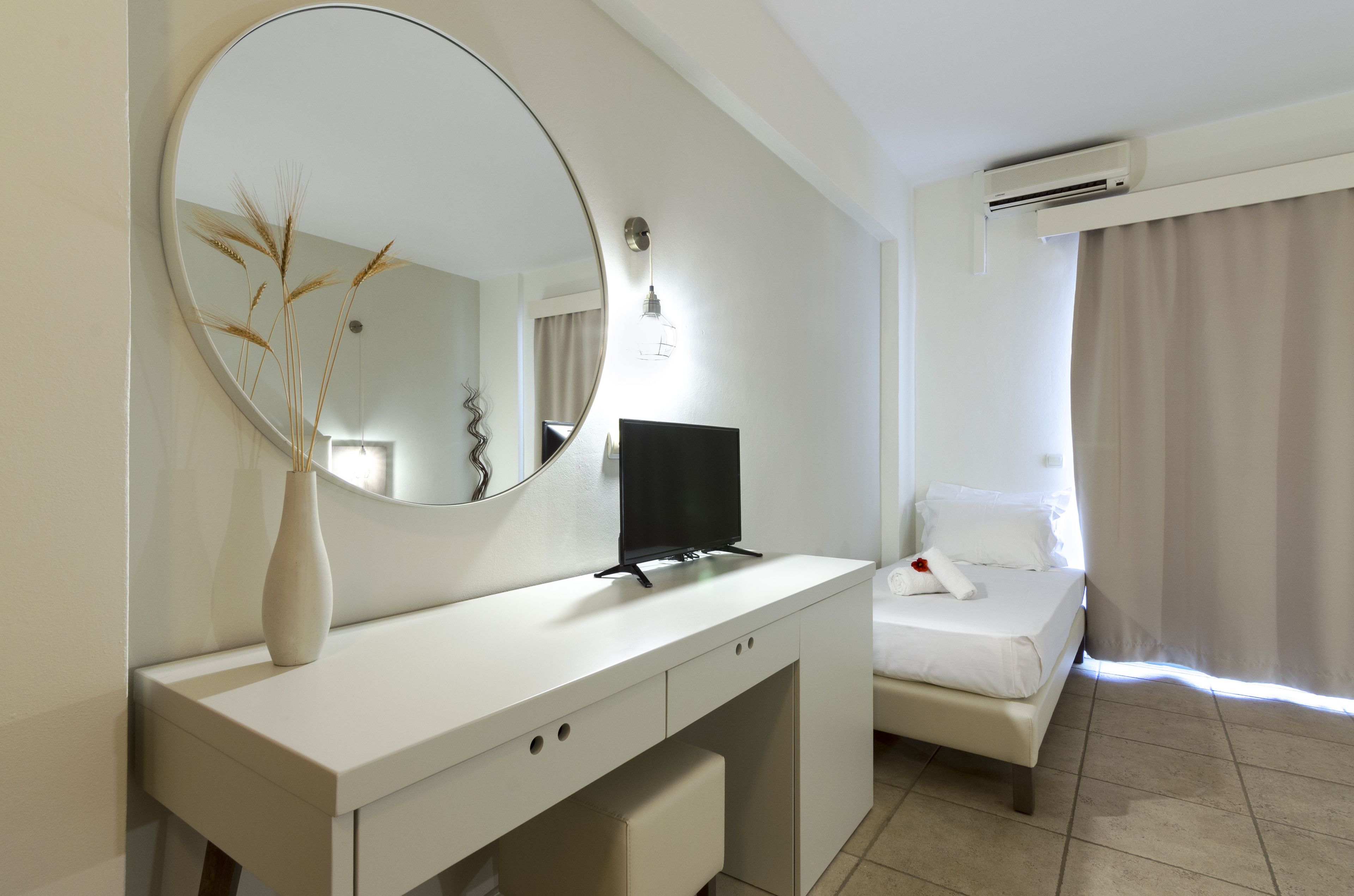 Habitación de hotel con cama blanca, televisor sobre un escritorio blanco, un espejo grande y un jarrón con plantas secas.