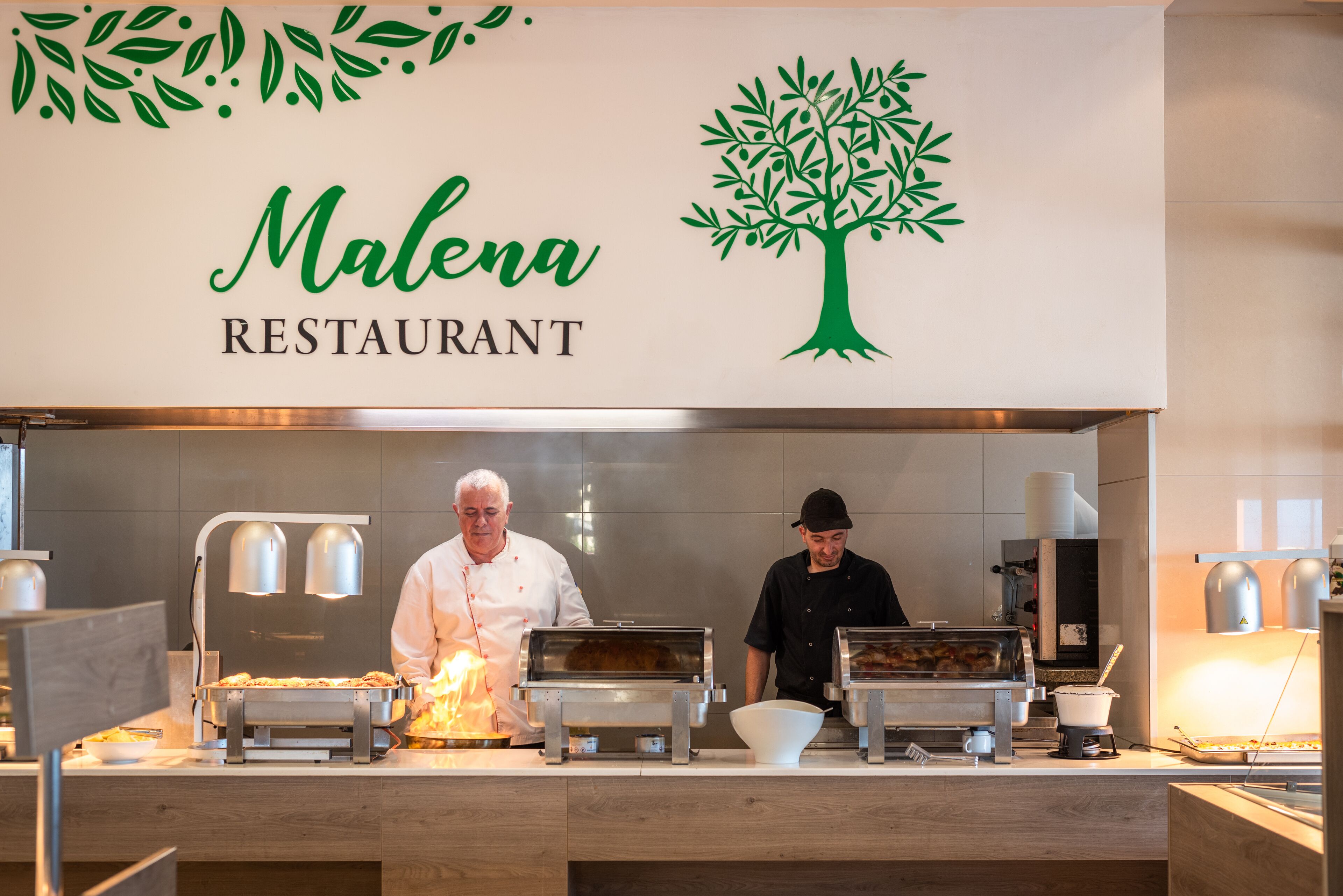 A restaurant counter with a sign reading "Malena Restaurant" and two chefs preparing food.