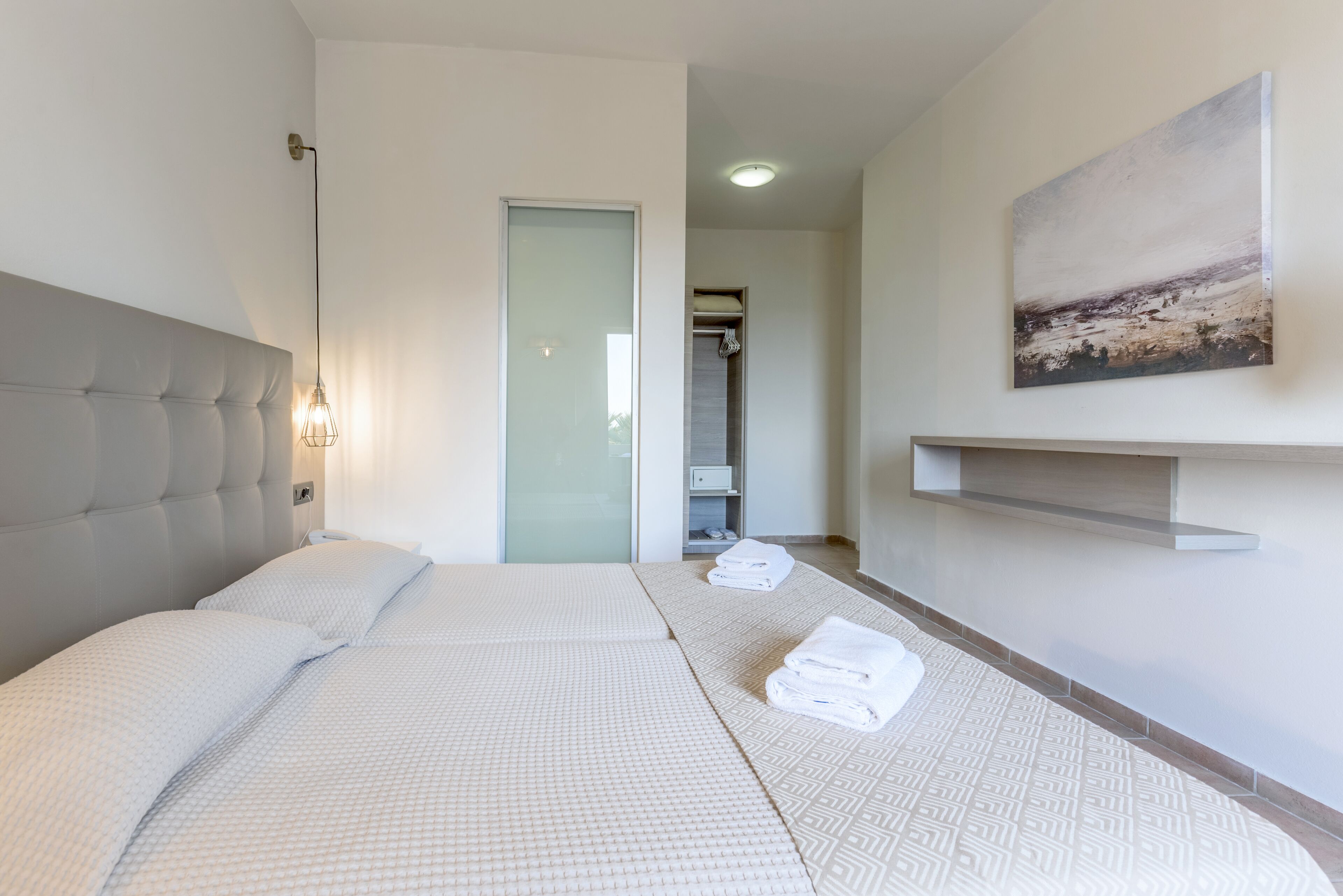 A hotel room with a large bed, a bedside lamp, a painting on the wall, and a bathroom visible through a glass door.