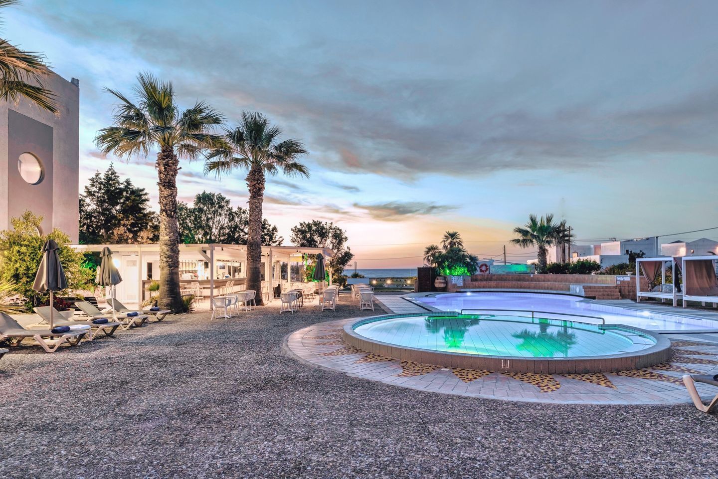 Zona de piscina amb gandules i ombrel·les, envoltada de palmeres i un edifici amb una finestra circular.