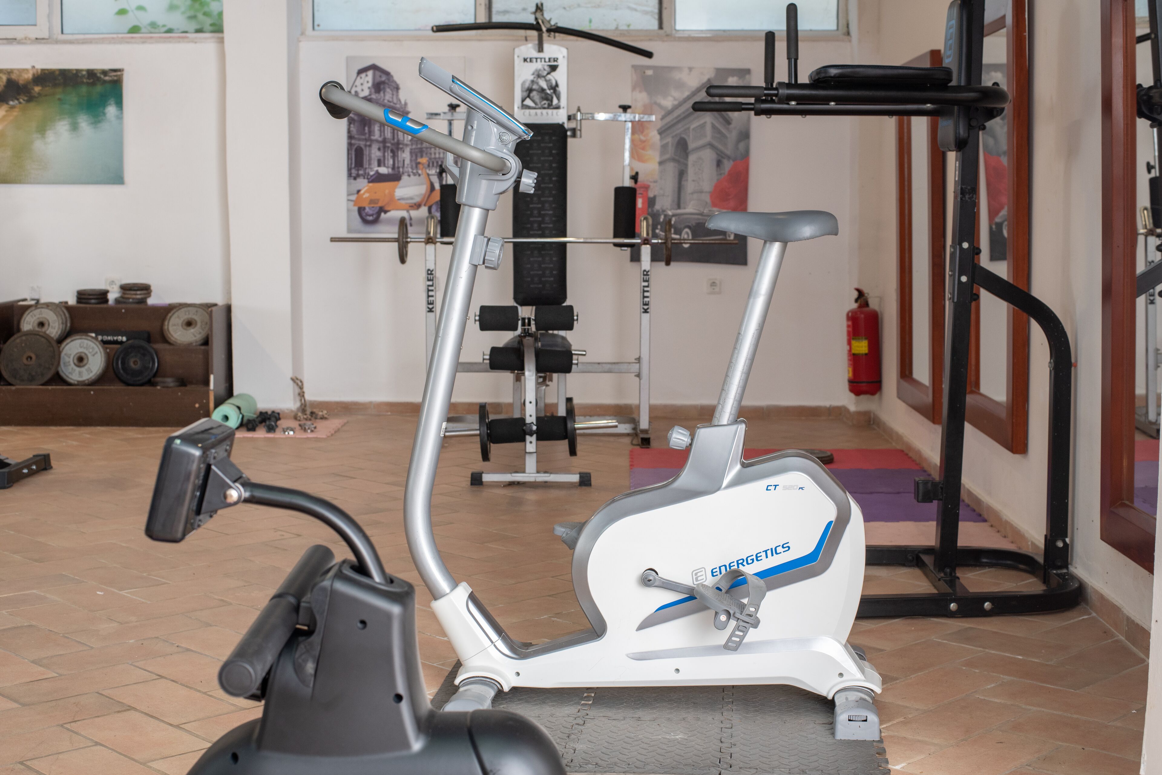 An exercise machine with a handlebar and a seat, a weight bench, and a wall-mounted rack with weights.