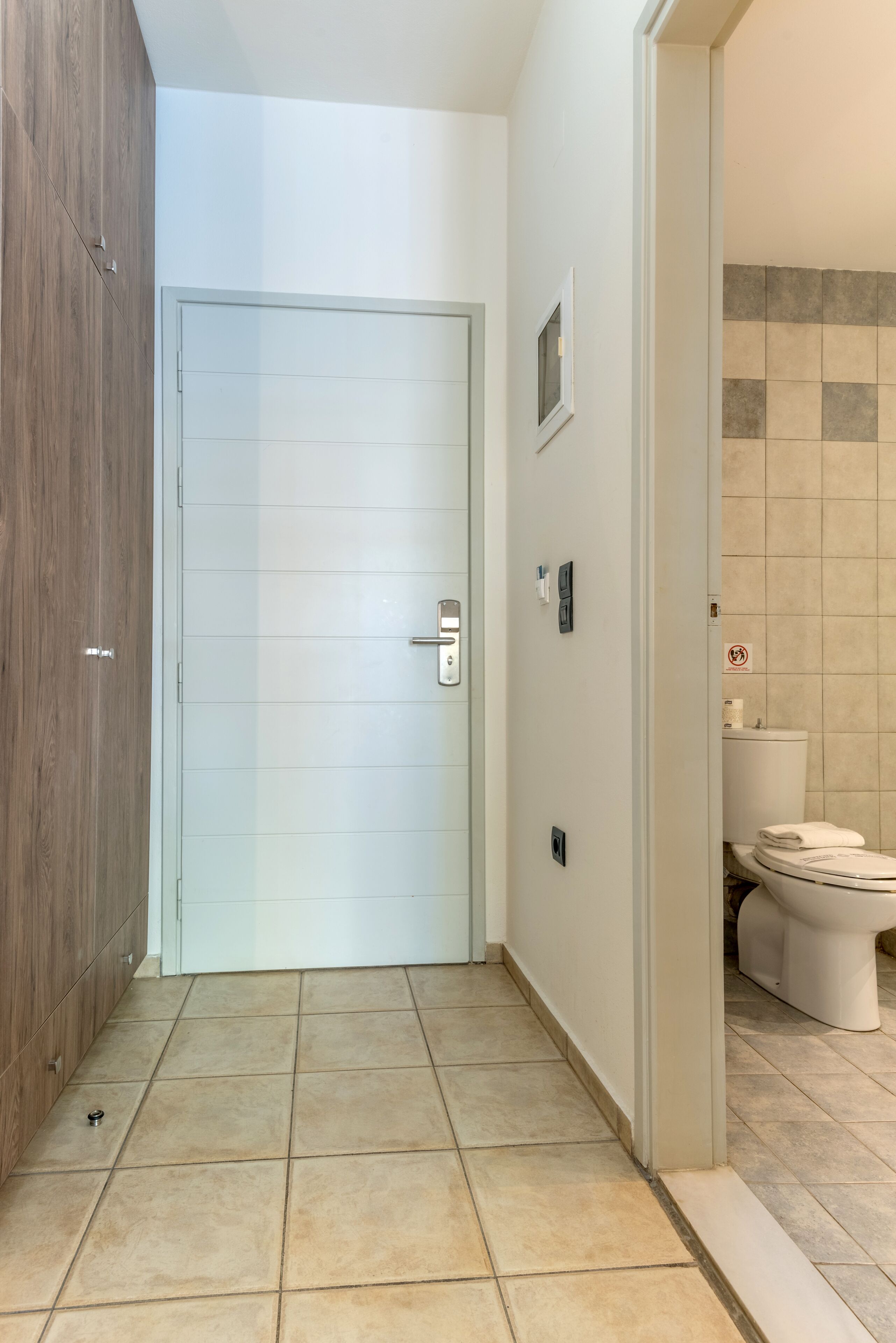 Um corredor com uma porta de madeira, uma porta branca e um banheiro com vaso sanitário e paredes azulejadas.