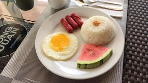 Un plato con huevo, arroz, salchichas y una tajada de sandía sobre una mesa con una taza y cubiertos.