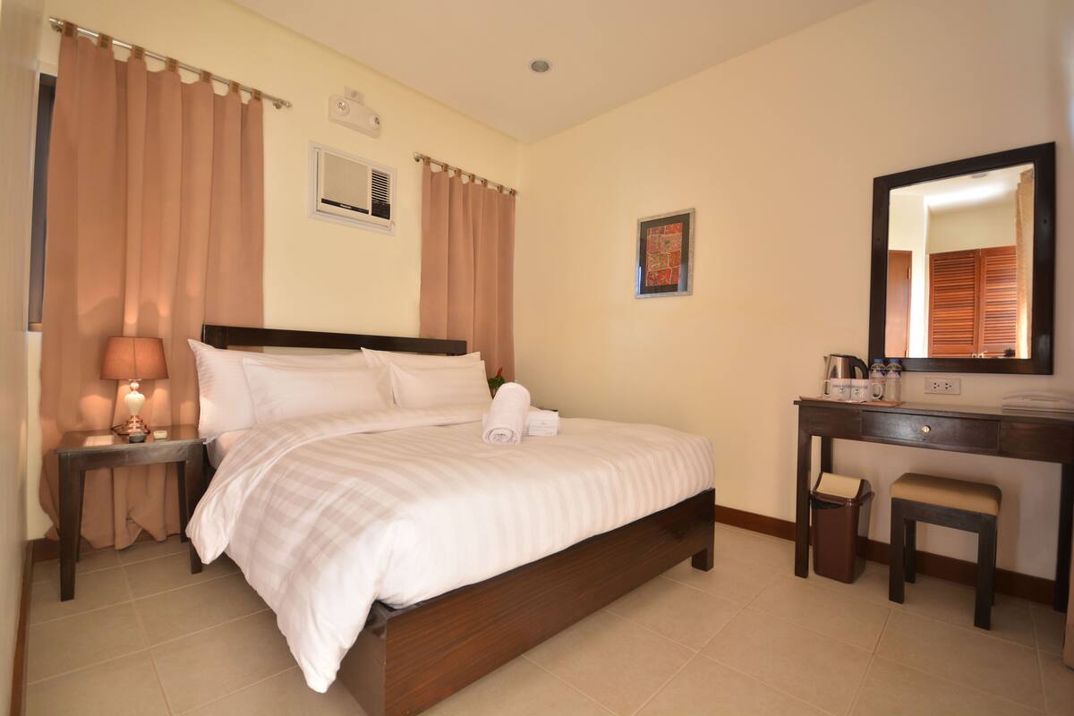 A hotel room with a bed, a desk, a chair, a lamp, a mirror, and a small trash bin.