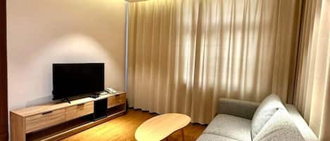 A modern living room with a flat-screen TV on a wooden stand, a grey sofa with cushions, a small wooden coffee table, and a large window with curtains.