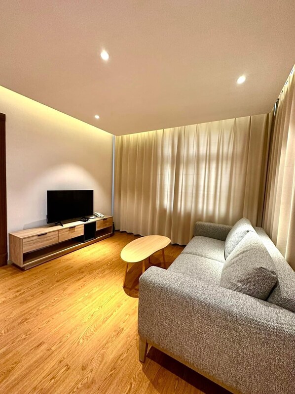 Una sala moderna con un televisor de pantalla plana sobre un soporte de madera, un sofá gris con cojines, una mesita de centro de madera y un ventanal con cortinas.