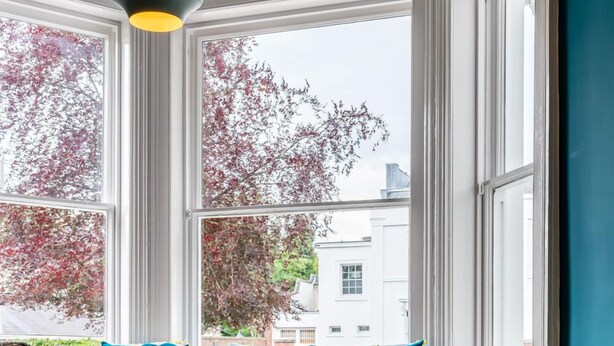 A cozy living room with a blue sofa, a wooden coffee table, and a patterned armchair. Large windows offer a view of trees and buildings outside. - The High Field Town House (Birmingham)