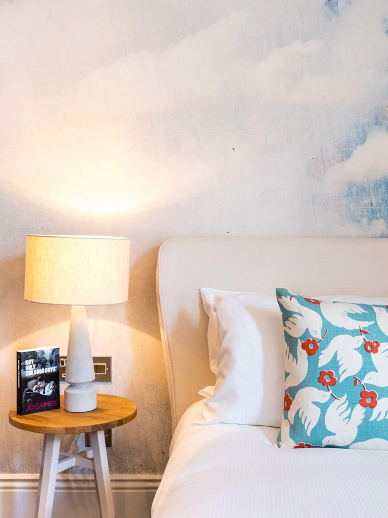 A neatly made bed with white linens, a floral pillow, and a bedside table with a lamp and a book.