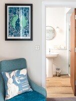 A room with a blue armchair, a framed artwork, a bathroom with a sink and mirror, and a wooden floor.