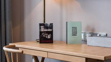 Ein Hotelzimmer mit einem Holztisch, einer Lampe, einem Buch mit dem Titel 'Not Only the Good Boys' von Jo Eames, einer grünen Karte und einem an der Wand befestigten Fernseher.