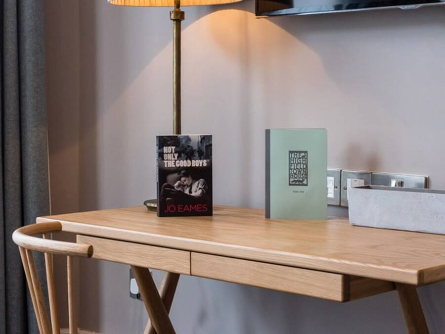Ein Hotelzimmer mit einem Holztisch, einer Lampe, einem Buch mit dem Titel 'Not Only the Good Boys' von Jo Eames, einer grünen Karte und einem an der Wand befestigten Fernseher.