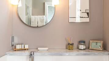 A bathroom with a round mirror, two wall-mounted lights, a sink, a framed picture, and a small shelf with toiletries.