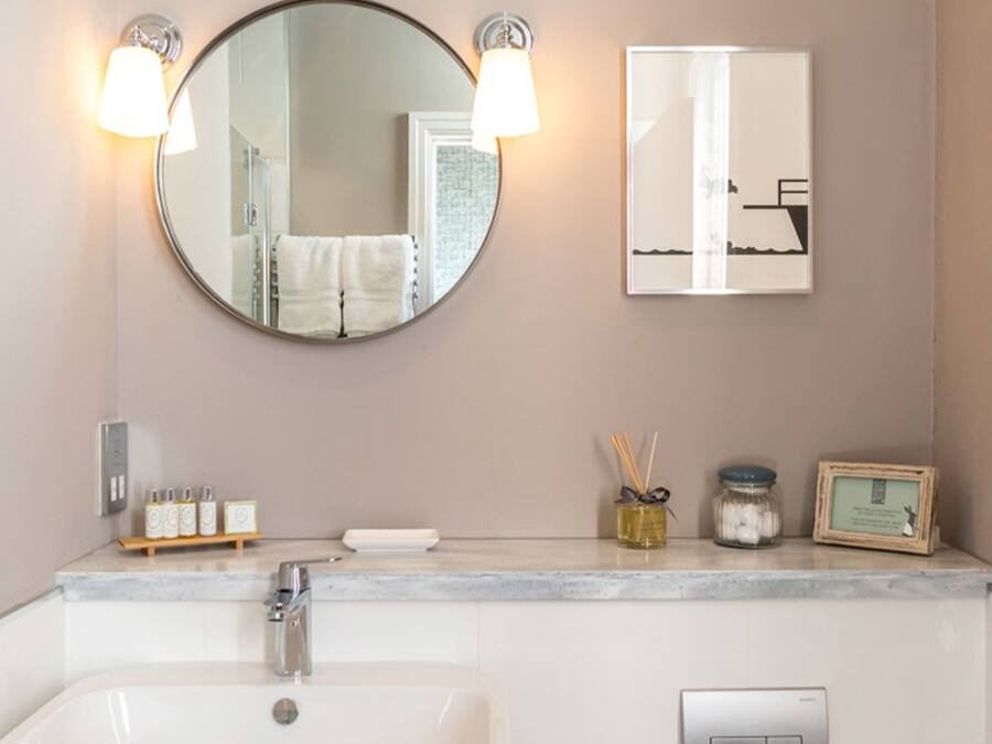A bathroom with a round mirror, two wall-mounted lights, a sink, a framed picture, and a small shelf with toiletries.