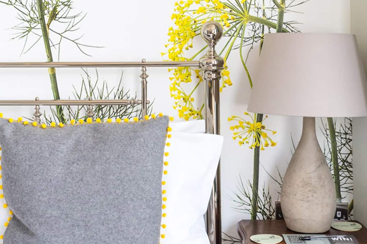 A neatly made bed with a grey pillow and a white pillow, a wooden nightstand with a lamp, and a wall with a floral wallpaper design.