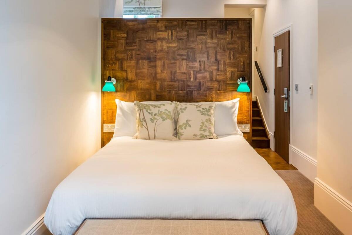 A bedroom with a wooden headboard, a bed with white bedding, two bedside lamps, and a staircase leading to another room.