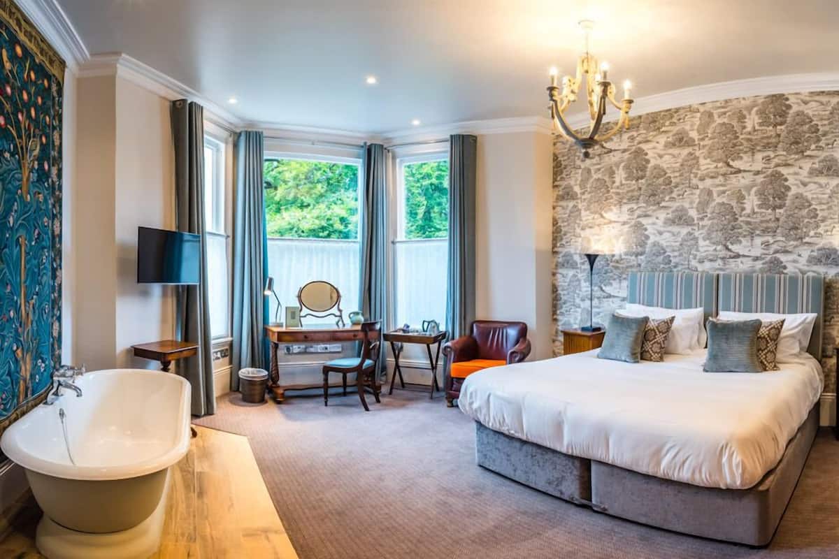 A bedroom with a large bed, a stone wall, a chandelier, a desk with a chair, a vanity with a mirror, and a bathtub.