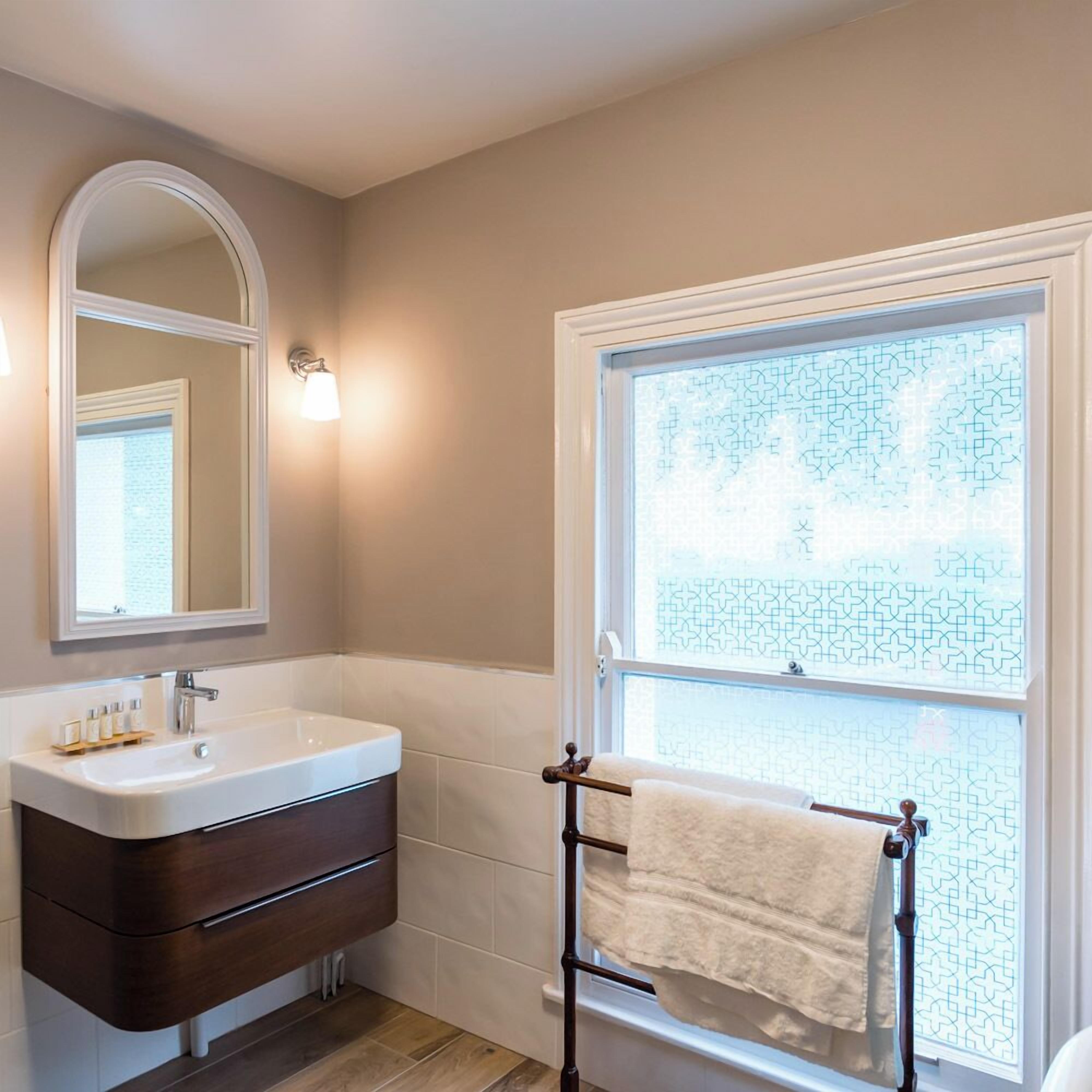 Un baño con lavamanos blanco, tocador de madera oscura, espejo y ventana con vidrio ornamental.