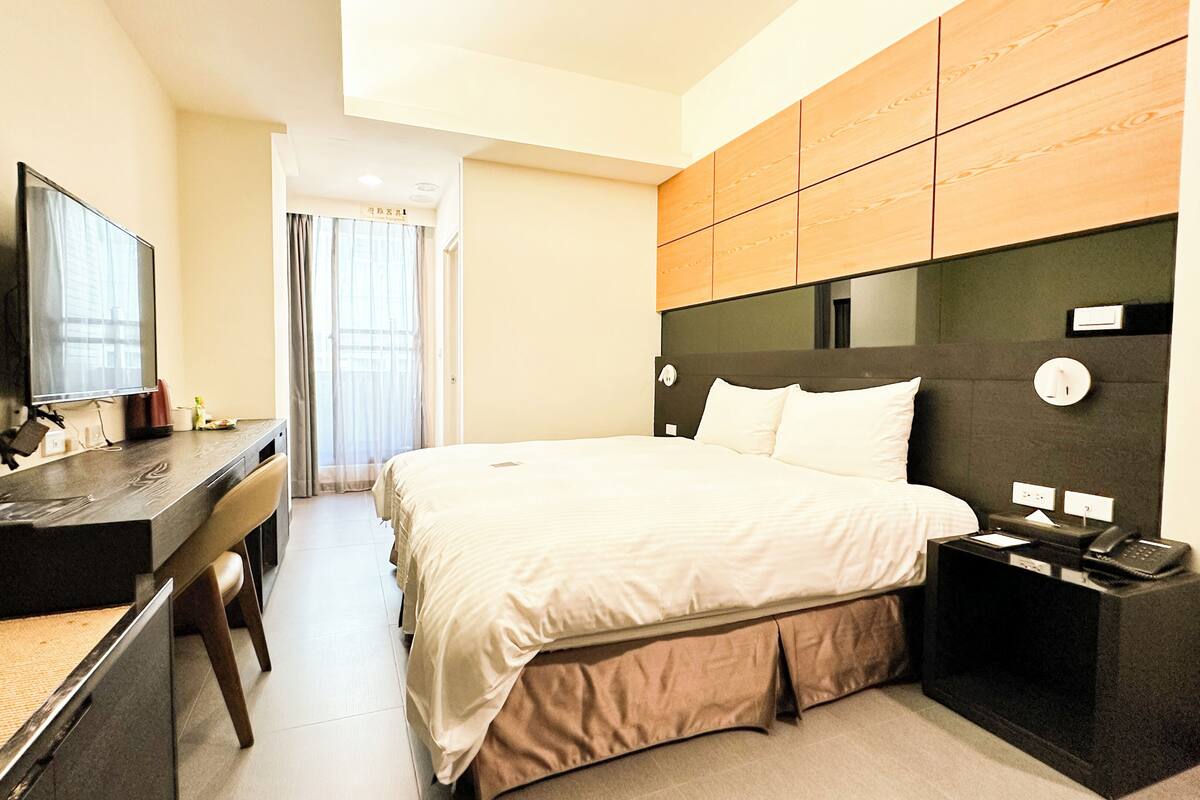 A hotel room with a bed, a desk with a TV, a telephone, and a plant.