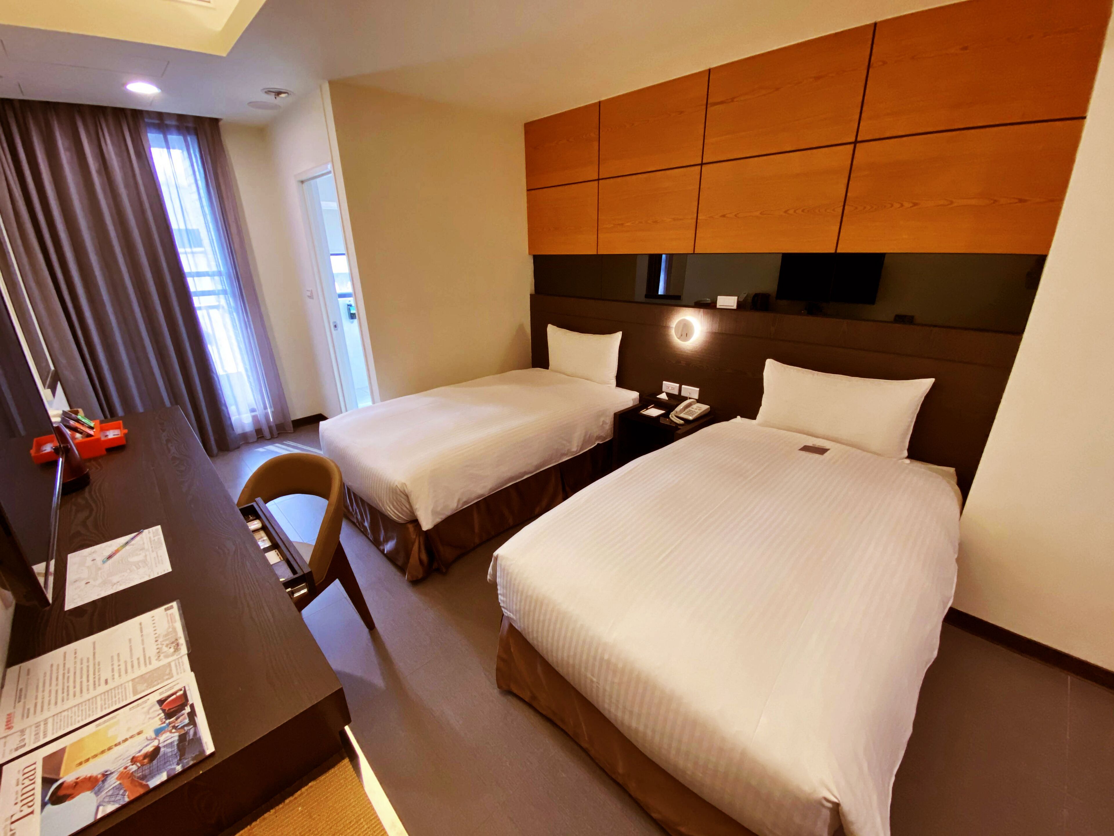 A hotel room with two beds, a desk, and a chair. There is a nightstand with a phone and a lamp.