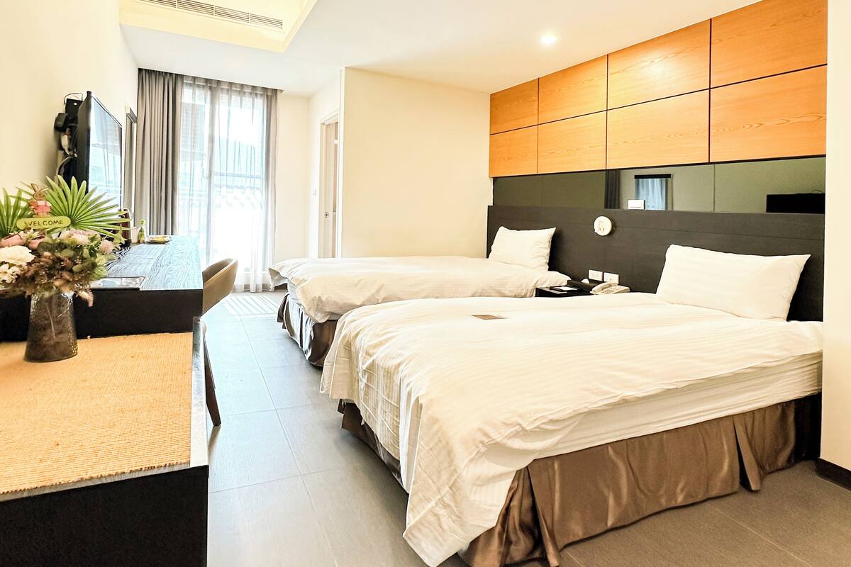 A hotel room with two beds, a desk, a chair, a vase with flowers, and a view of the outside.