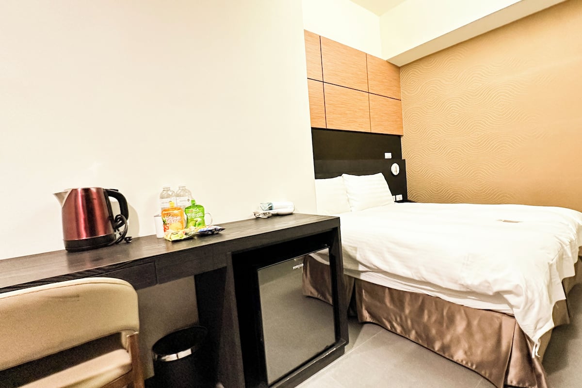 A hotel room with a bed, a desk with a kettle, and a trash bin.
