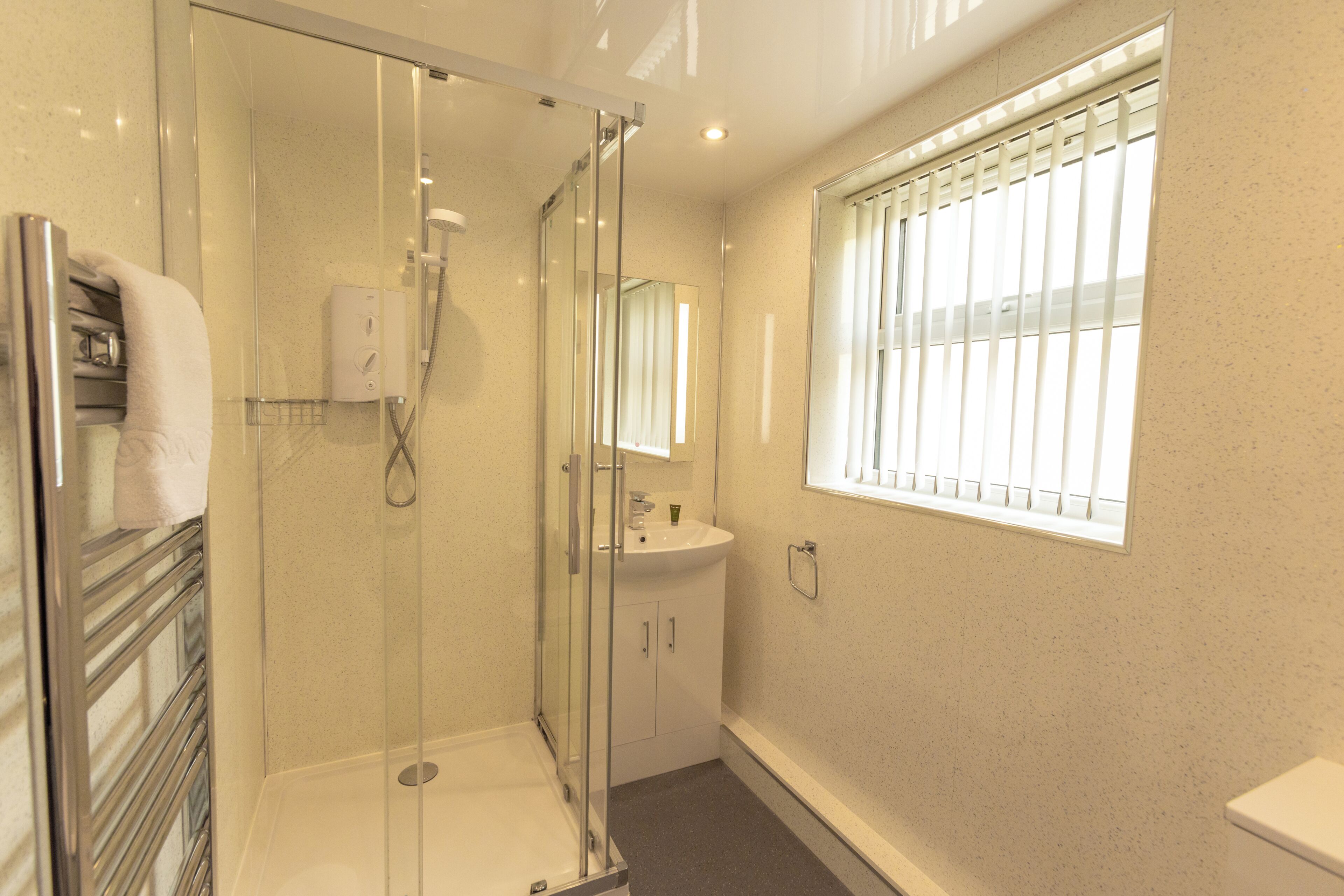Ein modernes Badezimmer mit einer gläsernen Duschkabine, einem weißen Waschbecken und einem Fenster mit Jalousien.