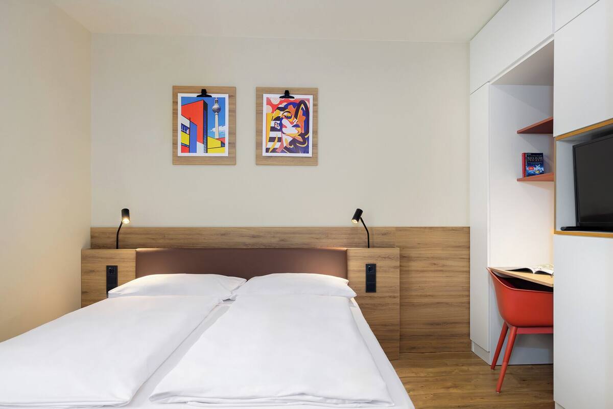 Una habitación de hotel con una cama, dos obras de arte colgadas en la pared, un escritorio con una silla y un televisor.