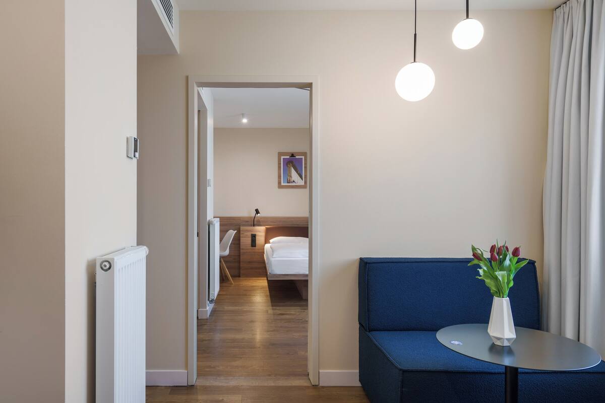Habitación de hotel con cama, mesita de noche con un jarrón de flores, un sofá azul y vista a otra habitación.