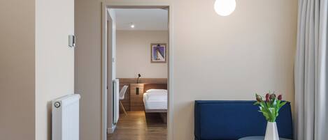 Habitación de hotel con cama, mesita de noche con un jarrón de flores, un sofá azul y vista a otra habitación.