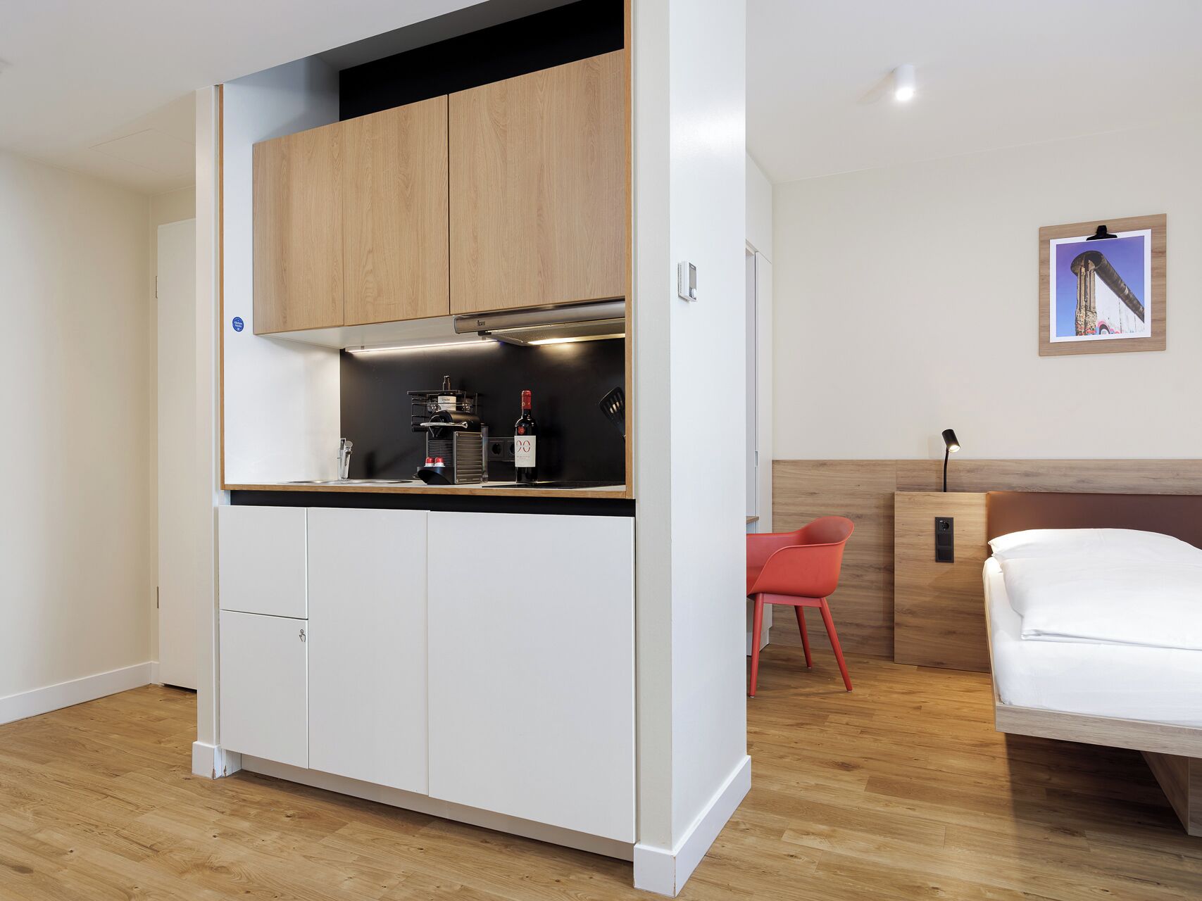 A compact room with a kitchenette, a bed, a red chair, and a framed picture on the wall.