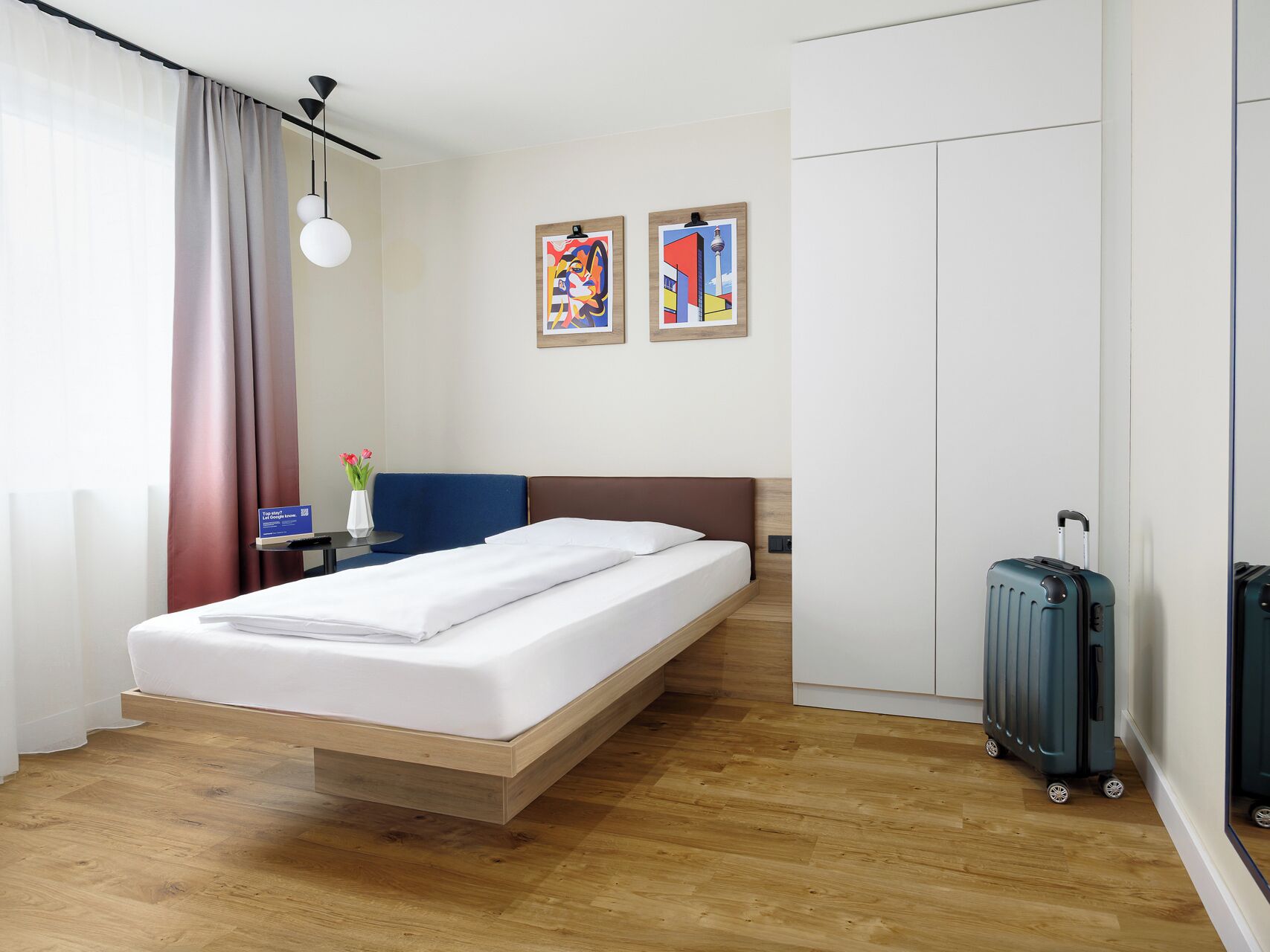 A hotel room with a bed, a desk, a chair, a suitcase, and two framed artworks on the wall.
